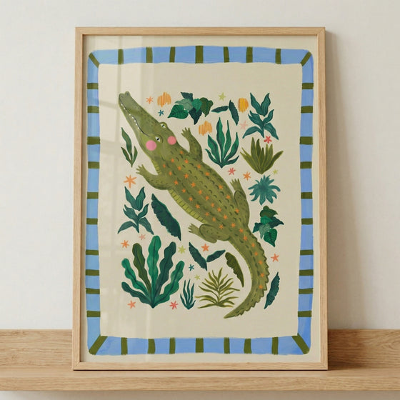 Framed illustration of a green crocodile with pink cheeks surrounded by leafy plants and small colorful flowers in a playful jungle style. The artwork sits on a wooden surface, showing how this whimsical animal print fits into nursery decor.