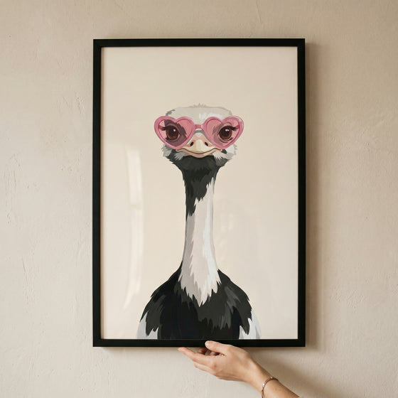 Framed pink heart glasses ostrich wall art held by a hand against a neutral wall.
