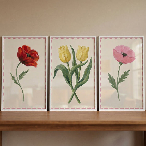 Three framed floral paintings with pink scalloped borders displayed side by side on a wooden surface featuring a red poppy yellow tulips and a soft pink bloom creating a warm balanced botanical arrangement.