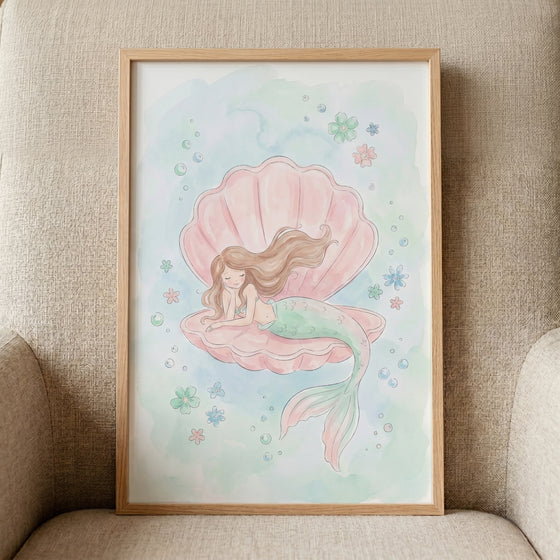 Framed pink shell sleeping mermaid wall art on a tan chair depicts a mermaid resting in a pink seashell with bubbles.
