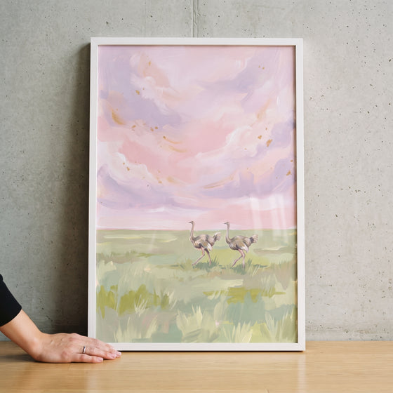 Pink sky walking ostrich animal wall art depicts two birds in a grassy field under soft pastel clouds in a white frame.
