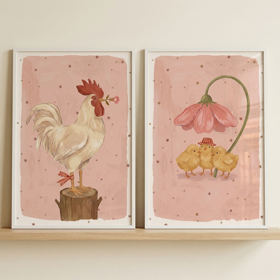 Two framed illustrations sit side-by-side on a light wooden shelf against a neutral wall. The left artwork features a white rooster on a tree stump, holding a pink flower in its beak with a red and white ribbon on its leg. The right artwork shows three yellow chicks under a large, drooping pink flower. One chick wears a small checkered hat. Both have soft pink backgrounds with tiny stars and are in white frames. The style is gentle and storybook-like.