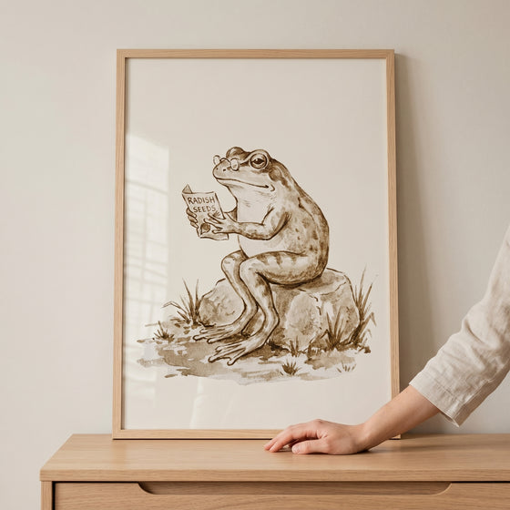 Framed reading radish seeds frog wall art shows a bespectacled frog sitting on a rock and reading a seed packet.