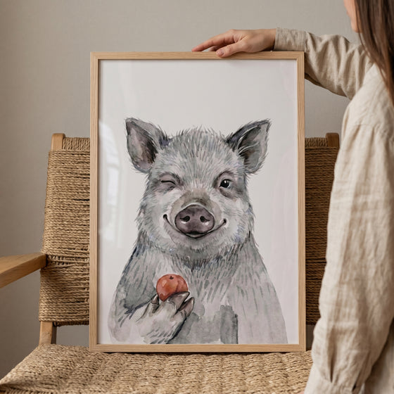 Framed red apple winking pig wall art resting on a woven chair, held by a person against a neutral wall.