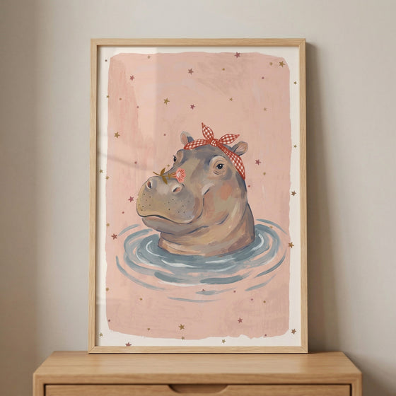 Framed illustration of a hippo floating in water with gentle ripples, wearing a red checkered headband and holding a small flower near its nose against a soft pink background with scattered stars. The playful artwork adds a calm and whimsical touch to a nursery or children’s room decor.