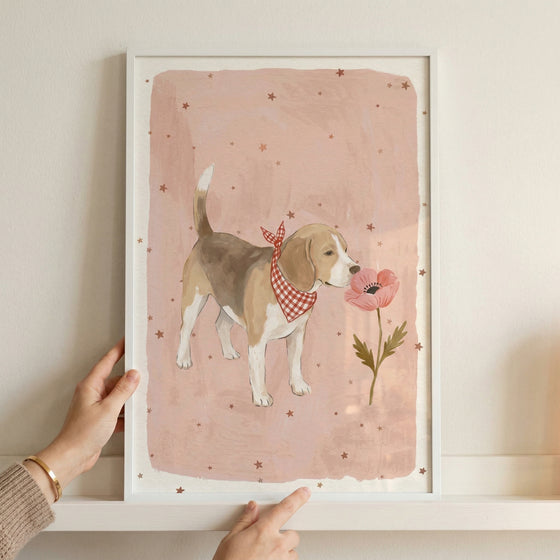 Framed illustration of a small brown and white dog wearing a red checkered bandana standing beside a pink flower on a soft blush background with scattered stars. A person’s hands hold the frame against a wall, showing the scale and adding a warm, styled nursery decor feel.