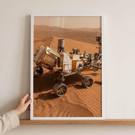 Framed red planet sand rover wall art depicts a robotic exploration vehicle on orange dunes, held by a hand on a shelf.