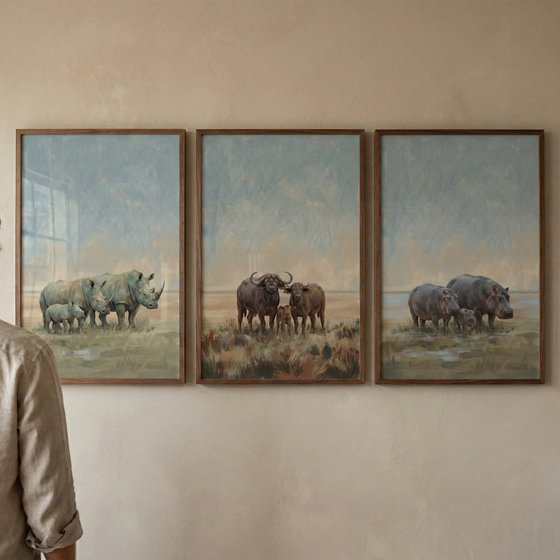 Three vertically-oriented framed artworks hang side-by-side on a light tan wall. Each piece is set in a thin, dark wood frame and depicts a different group of African safari animals in a muted, hazy style with soft blue skies. The frame on the left shows a rhinoceros family with a calf. The middle frame features three cape buffalo standing in tall, dry grass. The frame on the right portrays a group of three hippopotamuses near a body of water.