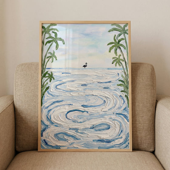 Framed painting of a calm ocean scene with textured swirling waves and a small bird standing near the shoreline framed by tall palm trees. The artwork rests against a cushioned chair highlighting its soft coastal colors and peaceful beach atmosphere.