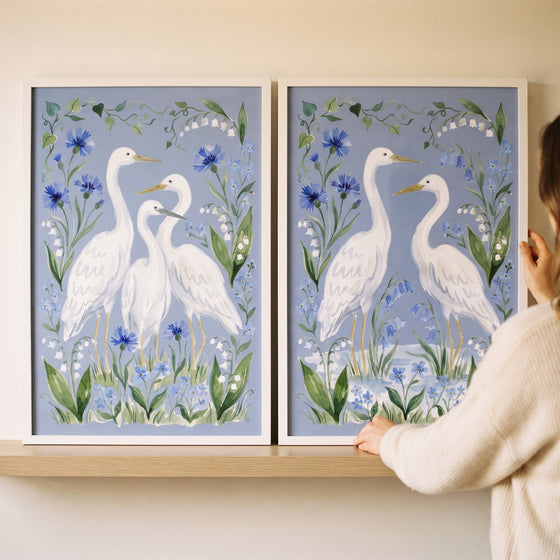 A woman adjusts a serene marshland white egret bird wall art set of 2 featuring blue flowers in white frames.