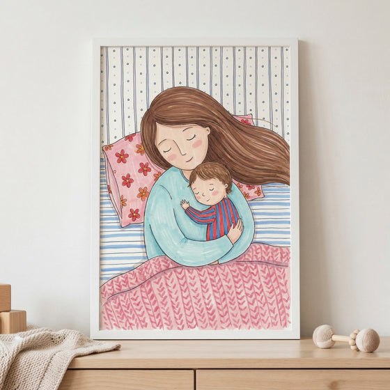 A framed illustration shows a mother and young child sleeping peacefully together. The mother has long brown hair and wears a light blue top, cradling the child who is in a red and blue striped shirt. They are in a bed with a pink flower-patterned pillow and a pink knit-patterned blanket. Behind them is a blue and white striped background with small dots. The white frame sits on a wooden shelf next to wooden blocks, a folded knit blanket, and a wooden rattle, creating a warm nursery setting.