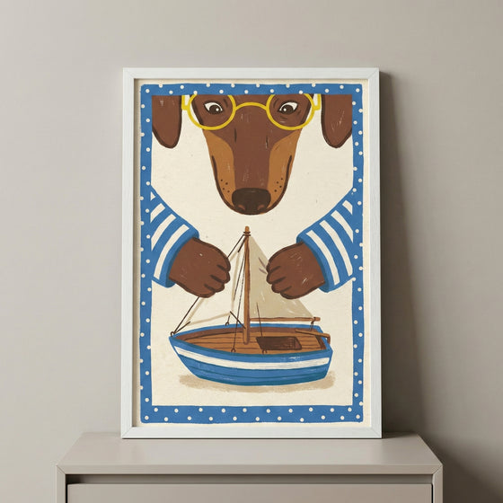A framed illustration features a brown dachshund with yellow glasses and a blue-and-white striped shirt. The dog appears to be carefully holding or building a small wooden sailboat with a white sail. The artwork is framed by a blue border with white polka dots and is displayed in a white frame on a light-colored surface against a neutral wall.