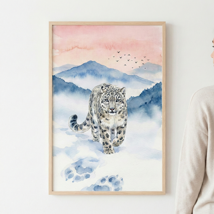 Vertical watercolor art featuring a majestic snow leopard walking forward through a snowy landscape. The animal has thick, spotted fur and is leaving large, blue-shadowed paw prints in the soft white snow. In the background, layers of misty blue mountains rise toward a sky filled with gentle pink and peach hues. A small flock of birds is silhouetted in the distance. The piece is held in a light wood frame and displayed on a neutral wall.