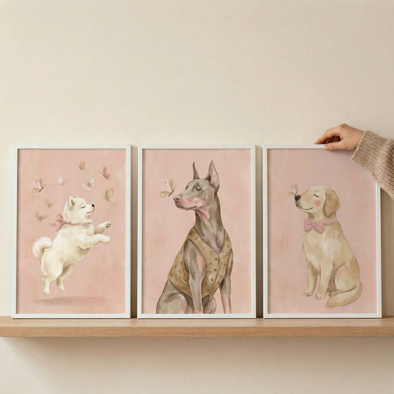 Three white-framed digital illustrations are displayed on a light wood shelf against a neutral wall. A person's hand, partially visible and wearing a beige knit sweater, is shown adjusting the frame on the right.