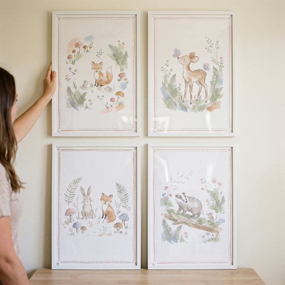 Person hangs a soft fern woodland animal wall art set of 4 featuring a fox, deer, rabbit, and badger in white frames.