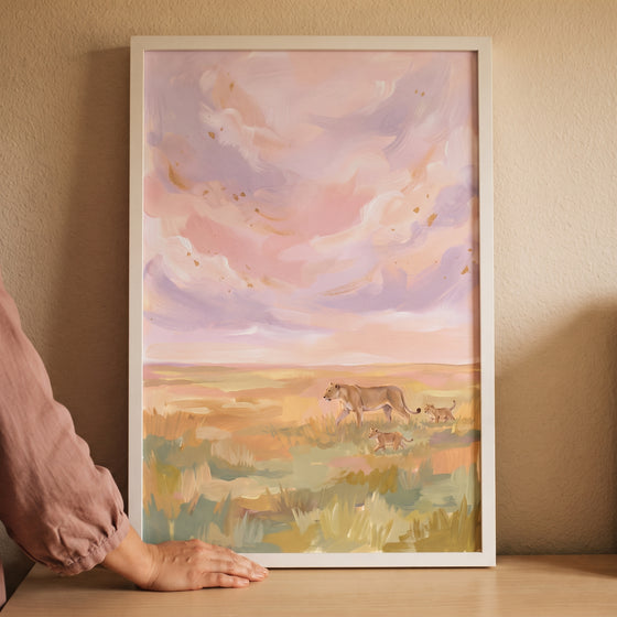 Soft lavender savanna lions wall art in a white frame touched by a hand, showing a lioness and cubs in a pastel field.