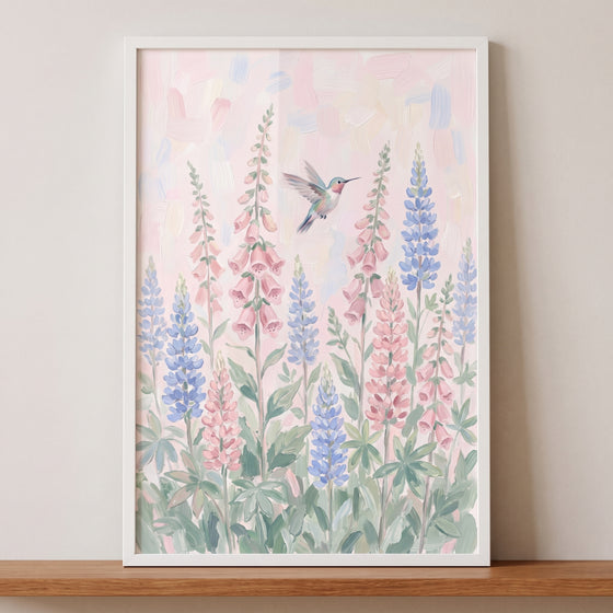Soft pastel flying hummingbird wall art featuring pink foxgloves and blue lupine flowers in a white frame.