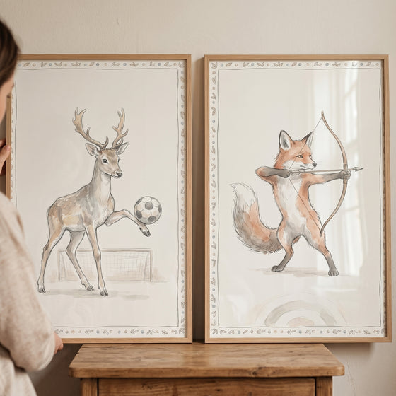 Soft watercolor athletic animals wall art set of 2: a deer plays soccer and a fox practices archery in wood frames.