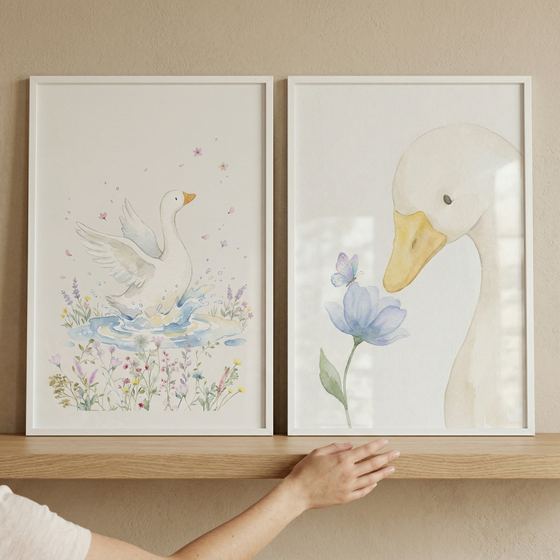 Two white-framed illustrations rest on a light wood shelf against a beige wall. The left art piece features a white goose in a small pond, surrounded by colorful wildflowers and floating pink petals. The right piece shows a close-up of a white goose looking down at a blue flower with a butterfly. Both pieces use a soft watercolor-style with pastel tones. A person's hand is shown reaching toward the shelf below the frames.