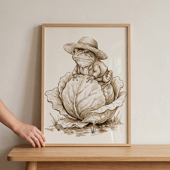 Straw hat garden frog wall art in a wooden frame shows a sepia illustration of a frog in a jacket sitting on a cabbage.