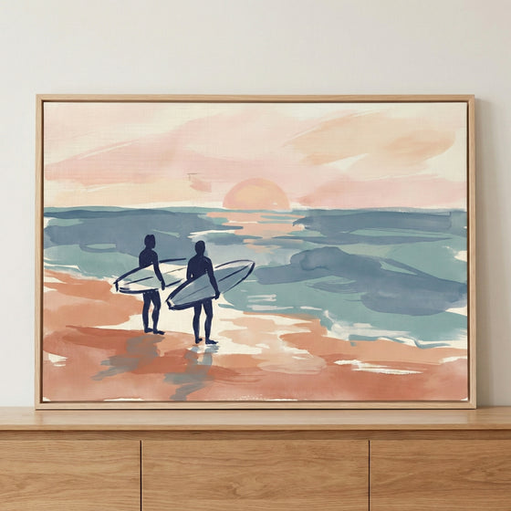 A framed illustration of two dark-blue silhouettes of surfers standing on a peach-colored beach, holding their surfboards and looking out at a calm, teal and blue ocean. A large, soft-peach sun sits low on the horizon in a pale pink sky. The style is soft and expressive with visible brushstrokes and a muted pastel color palette.