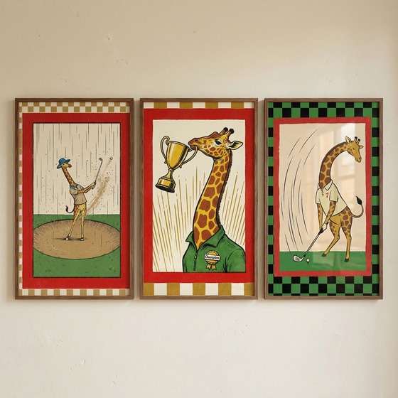 Three framed illustrations of giraffes playing golf are hung side-by-side on a plain wall.
The left illustration shows a giraffe wearing a small hat and a tan vest swinging a golf club in a sand trap. It has a red border and a tan-and-white checkered frame.