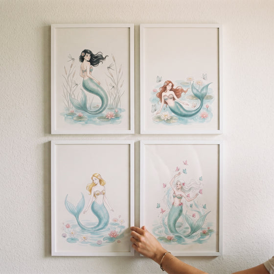 Teal tail watercolor mermaid wall art set of 4 in white frames being adjusted by a hand on a plain wall.