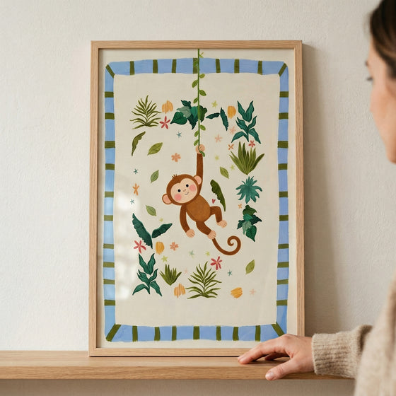 A framed illustration shows a cheerful brown cartoon monkey swinging from a green vine. The smiling monkey is surrounded by stylized jungle leaves and small colorful flowers on a cream background. A light blue border with green stripes surrounds the artwork inside a light wood frame. The frame sits on a wooden shelf against a plain wall, with a person's hand visible on the right side adjusting its position.