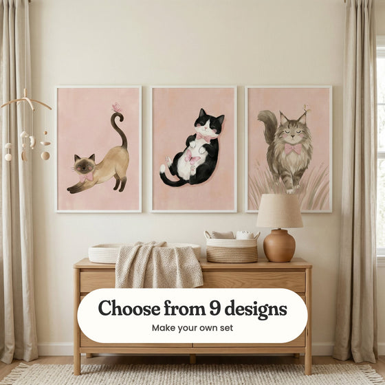 Nursery with three cat prints in white frames between linen curtains, above a light oak dresser with a knit throw, woven baskets and a terracotta lamp, beside a wooden baby mobile: Dainty Morning Stretch, Dreamy Butterfly Nap and Blushing Bow Feline, with a label reading Choose from 9 designs, Make your own set