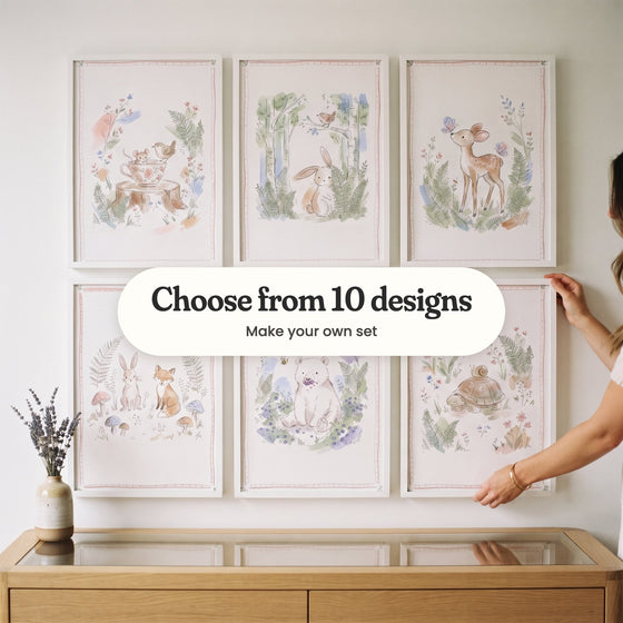 A hand straightening one of six woodland animal prints in white frames in two rows on a white wall above a light wood sideboard with a vase of lavender: a mouse in a flowered teacup with a wren, a bunny among birch trees, a fawn with butterflies, a bunny and a fox among mushrooms, a bear with blueberries and a tortoise with a snail on its shell, with a label reading Choose from 10 designs, Make your own set