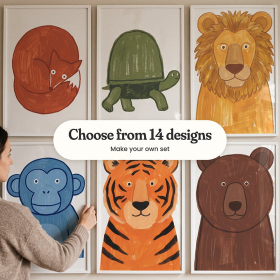Six bold animal prints in white frames in two rows on a beige wall, with a woman hanging the lower left one: a curled-up red fox, a green tortoise, a golden lion, a blue chimp, an orange tiger and a brown bear, with a label reading Choose from 14 designs, Make your own set