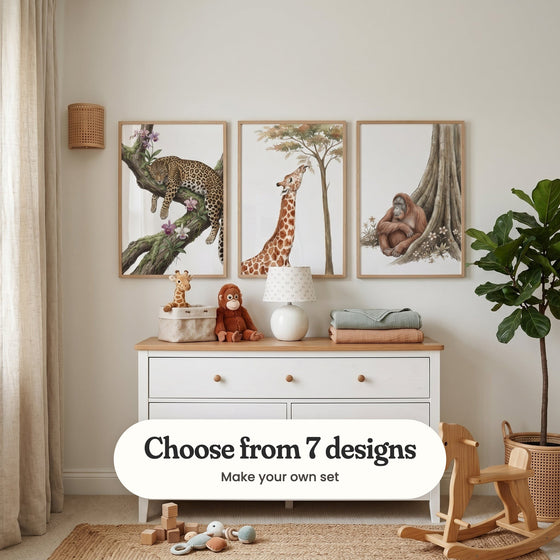 Cream nursery with three animal prints in light wood frames above a white dresser with a plush giraffe, a plush orangutan, a lamp and folded blankets, beside a fiddle leaf fig and a wooden rocking horse: Spotted Floral Repose, Savanna Snack Time and Gentle Jungle Ape, with a label reading Choose from 7 designs, Make your own set