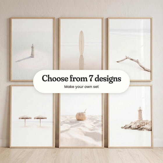 Six beach prints in light wood frames against a cream wall, three hanging on the wall above three leaning on a pale wood floor: a small white lighthouse on a sandbar, a wooden surfboard standing in shallow water, a bleached driftwood branch on the sand, two straw umbrellas on white sand, a coconut with a straw and a lighthouse on a rocky outcrop, with a label reading Choose from 7 designs, Make your own set