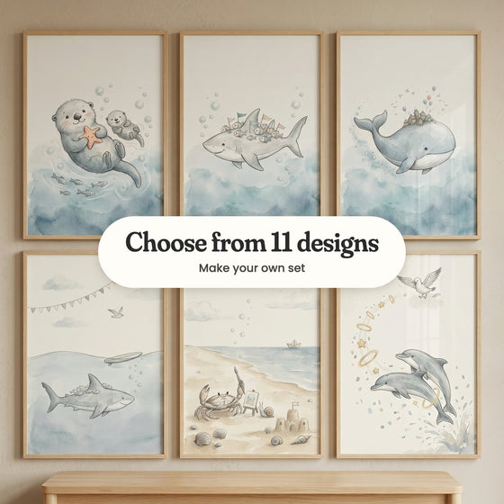Six sea animal prints in light wood frames in two rows above a light wood bench: a sea otter hugging a starfish beside its baby, a shark carrying fish with little flags, a whale with a tiny village on its back, a shark swimming under a surfboard, a crab holding a paintbrush at a little easel on the beach and two dolphins leaping through gold rings, with a label reading Choose from 11 designs, Make your own set