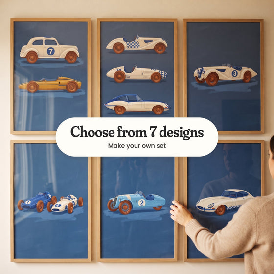 Six vintage race car prints on navy in light wood frames on a wall while a woman straightens one, with a label reading Choose from 7 designs, Make your own set