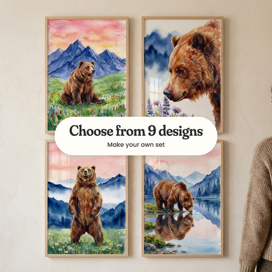 Four bear prints in light wood frames in two rows on a cream wall, with a woman in a brown knit sweater at the right: a bear sitting in a wildflower meadow under purple peaks, a close-up bear sniffing purple lupines, a bear standing on its hind legs in a meadow and a bear drinking from a still lake, with a label reading Choose from 9 designs, Make your own set