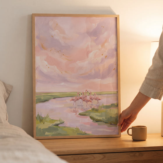 Wading blush pink flamingo animal wall art in a wood frame on a nightstand depicts birds in a marsh under a pastel sky.