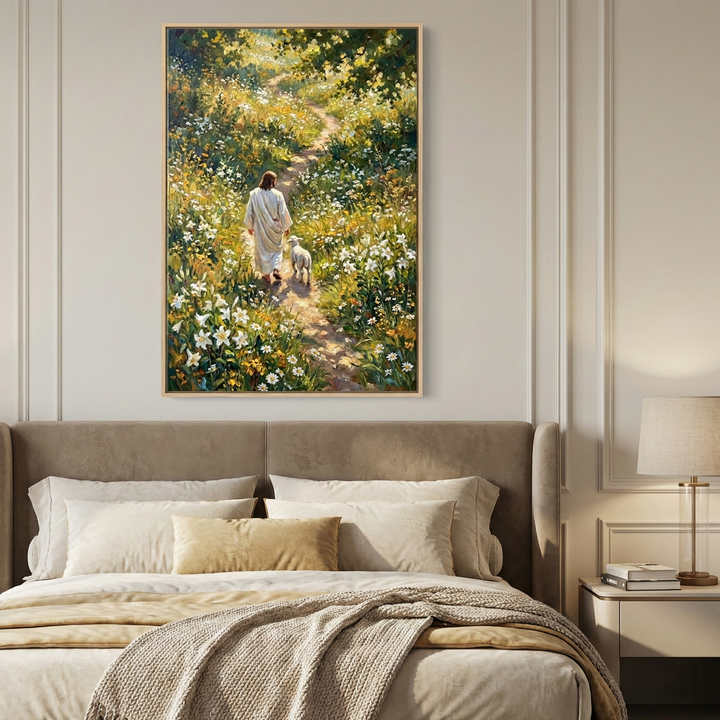 A bedroom features a large artwork centered above an upholstered tan headboard. The artwork depicts Jesus in a white robe walking along a sun-dappled dirt path through a field of vibrant wildflowers, accompanied by a small white lamb. The bed is layered with soft neutral-toned pillows and textured blankets. To the right, a nightstand holds a stack of books and a lamp with a cylindrical base and beige shade. The walls are light-colored with elegant decorative molding.