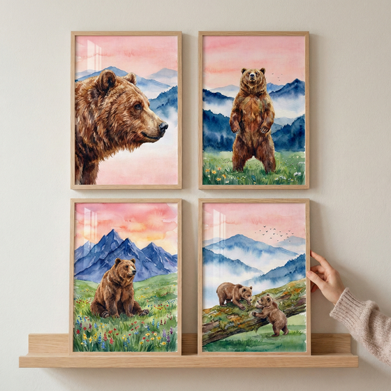 A set of four framed watercolor-style illustrations is displayed in a two-by-two grid on a light wooden shelf against a neutral-colored wall. Each artwork features a brown bear in various poses within a scenic mountain landscape under a soft pink and orange sky. The top-left piece shows a close-up profile of a bear's head against blue mountain peaks. The top-right image depicts a bear standing on its hind legs in a grassy field dotted with yellow flowers.