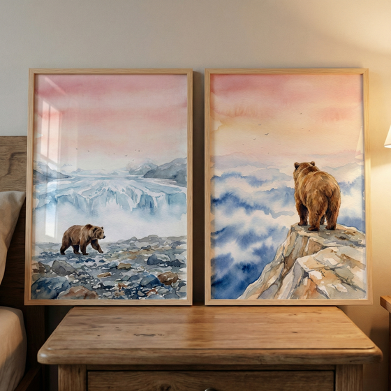 Two framed watercolor-style artworks sit on a wooden nightstand. In the left piece, a brown bear walks across a grey, rocky foreground with an icy blue landscape and a pastel pink sky behind it. The right piece features a bear seen from behind, standing on the edge of a rocky mountain peak looking out over a thick layer of blue and white clouds under a soft, warm sky. Both images are housed in light-colored wood frames and leaning against a neutral wall next to a bed.