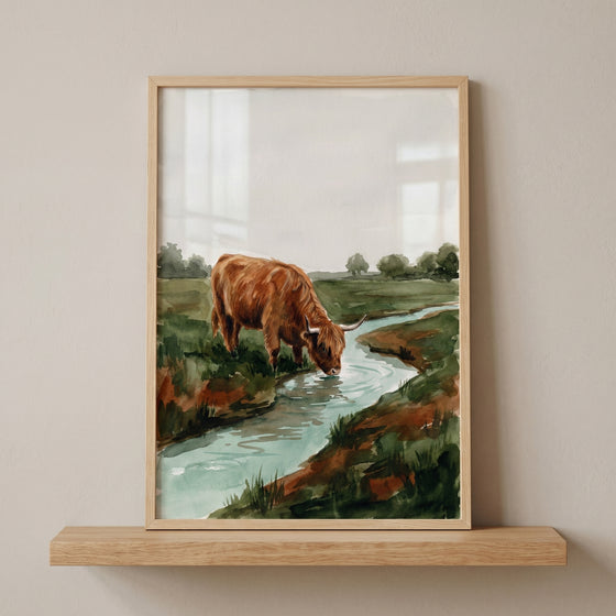 Framed watercolor drinking highland cow wall art on a shelf shows a brown cow by a stream in a green field.