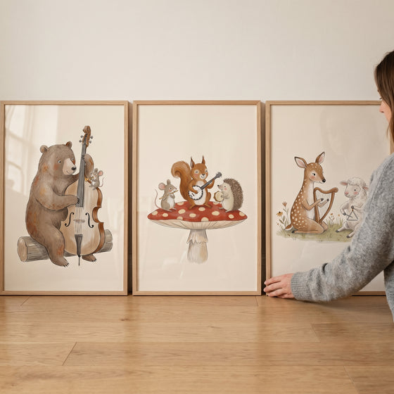 Watercolor musical woodland animal wall art set of 3 in wooden frames leaning against a wall on a light wood floor.