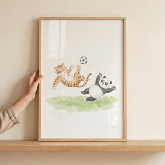 Watercolor soccer animals wall art depicts a tiger and panda playing soccer, in a wooden frame held by a hand.