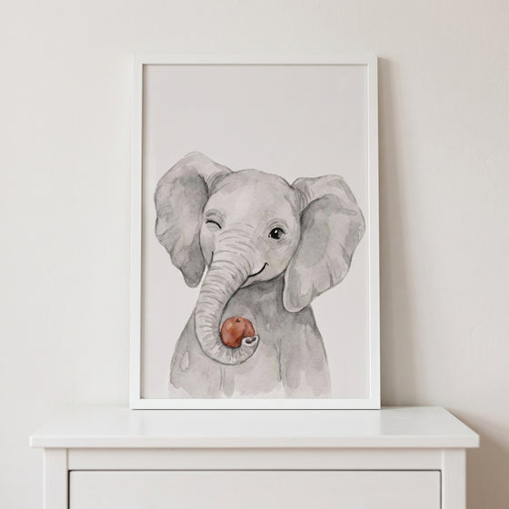Framed watercolor winking elephant wall art depicts a grey baby elephant holding a red apple with its trunk.