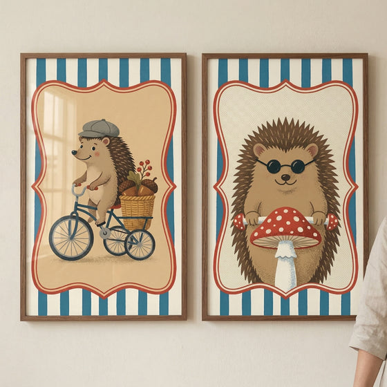 Two framed illustrations of anthropomorphic hedgehogs hang side-by-side on a light, neutral-toned wall. Both pieces feature a central character within a decorative, scalloped red frame, set against a background of vertical blue and white stripes. In the frame on the left, a hedgehog wearing a grey flat cap is depicted riding a blue tricycle. A small wicker basket on the back of the tricycle is filled with acorns and a sprig of red berries. The interior background behind the character is a light tan color.