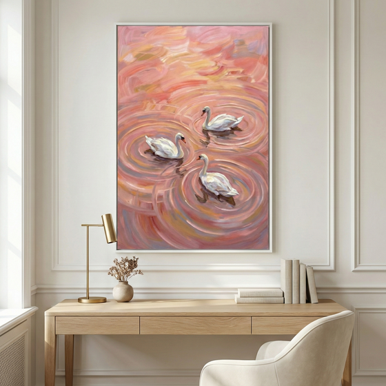 An interior view of a serene room featuring a large framed vertical canvas hanging on a paneled wall. The artwork depicts three elegant white swans floating in a sea of vibrant pink, rose, and peach-colored ripples, created with bold, expressive brushwork. Below the wall decor is a light wood desk with a clean design. The desk's surface holds a slender brass lamp on the left, a small neutral-toned vase with delicate dried sprigs, and several books in white and gray covers arranged neatly on the right.