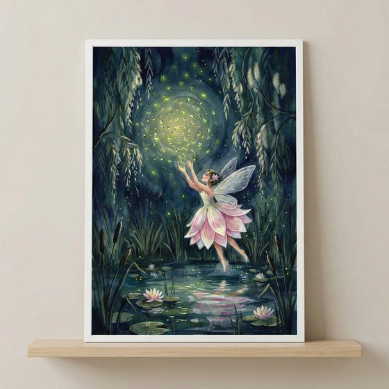 A framed piece of artwork rests on a light wood shelf against a neutral-colored wall. The illustration depicts a small winged figure in a dress that looks like soft pink flower petals. She is hovering above a dark pond at night, surrounded by lily pads and blooming white water lilies. Her arms are reaching upward toward a luminous, swirling cloud of glowing yellow fireflies. The scene is framed by dark green weeping branches and tall reeds, creating a magical and ethereal atmosphere.