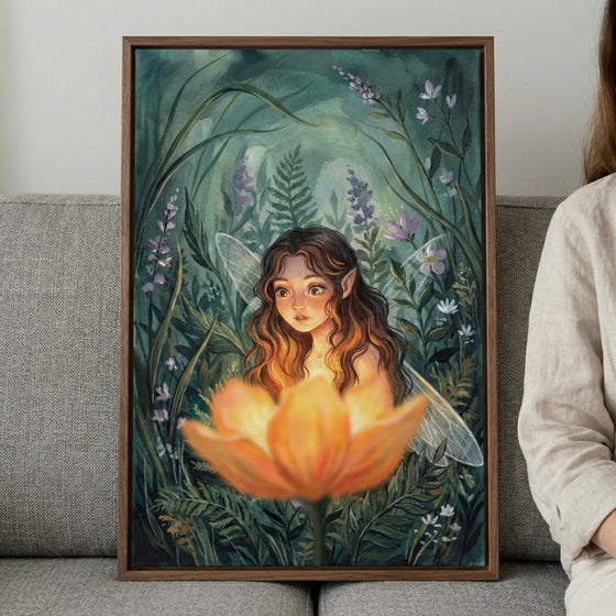 A framed illustration of a young fairy with pointed ears and long, wavy brown hair looking down at a large, glowing orange flower. The fairy is surrounded by tall blades of grass, ferns, and small purple and white flowers in a dark, ethereal forest setting. The glow from the flower lights up her face and hair. The framed art piece is resting on a gray couch.