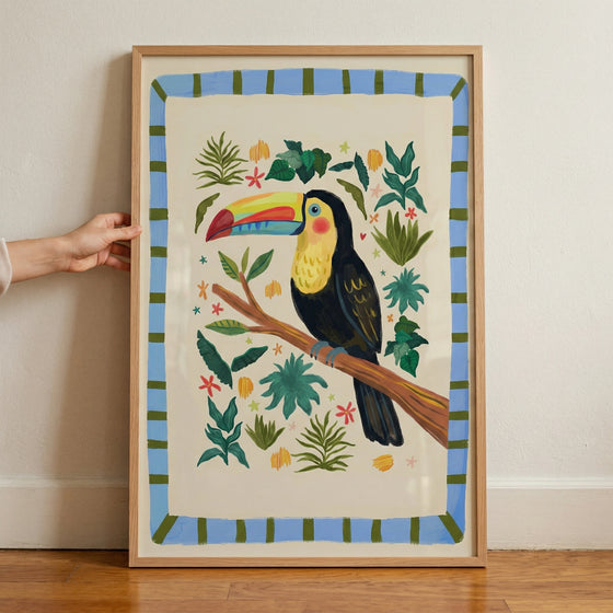 Framed illustration of a colorful toucan perched on a branch surrounded by green leaves and small bright flowers, set against a soft cream background with a blue and green striped border. A hand is visible holding the frame, adding context to the vibrant and playful jungle themed artwork.