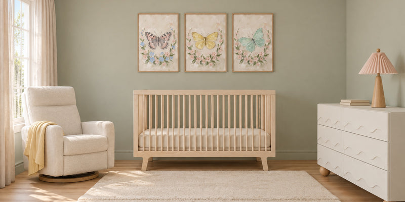 Butterfly nursery: three framed butterfly wreath prints in blush, butter yellow and mint above a pale birch crib, with sage walls, a cream glider and a white wave dresser